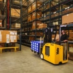 A Yale forklift transporting goods through narrow aisles of pallet rack in a warehouse.
