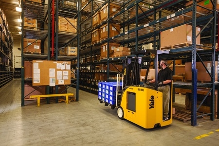 A Yale forklift transporting goods through narrow aisles of pallet rack in a warehouse.