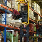 A pair of forklift operators retrieving goods from pallets high up on a shelf in a warehouse.