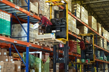 A pair of forklift operators retrieving goods from pallets high up on a shelf in a warehouse.